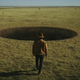 A man dressed in cowboy attire strides toward a giant hole in the ground.